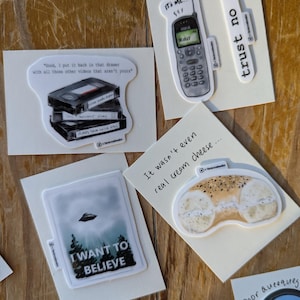 May include: A collection of stickers on a wooden surface. The stickers feature various designs, including a retro cell phone, a stack of VHS tapes, a UFO scene with the text "I WANT TO BELIEVE", and a bagel. Some stickers have text like "trust no one".