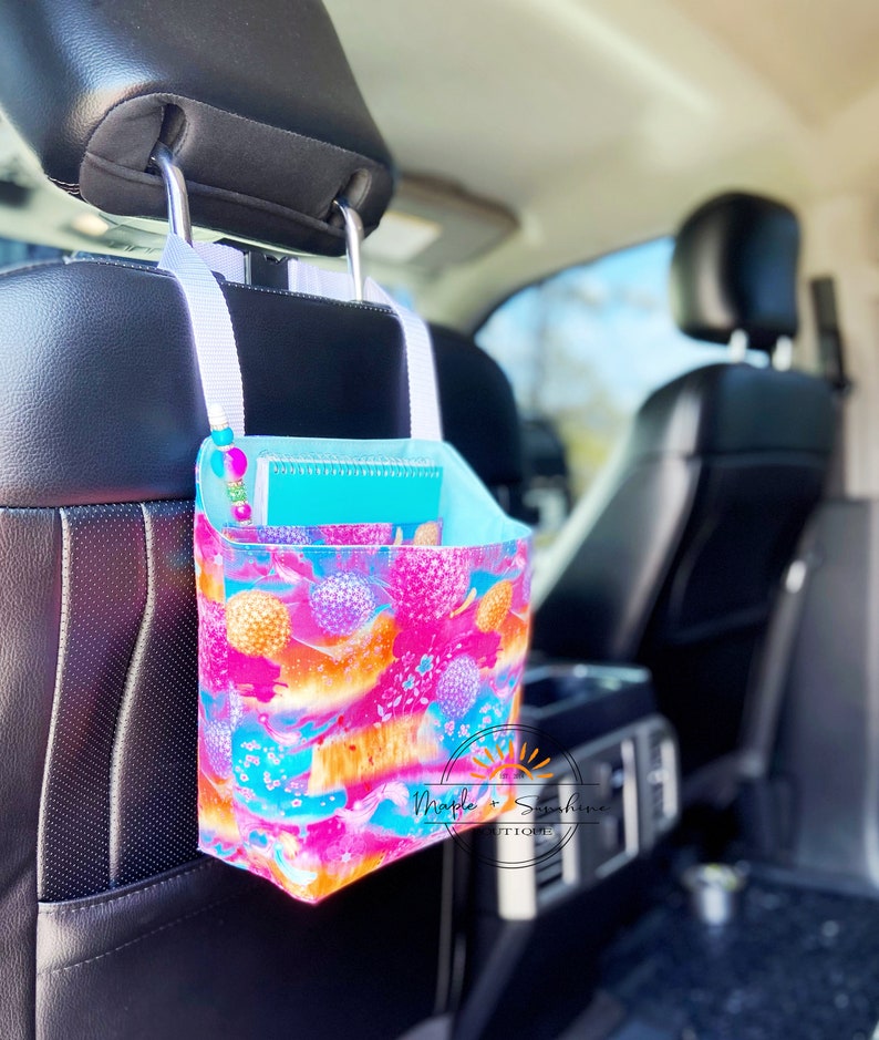 May include: A pink, blue and orange patterned fabric car organiser hanging from the headrest of a car seat. The organiser has a notebook and a pen inside. The text "Maple + Sunshine Boutique" is visible on the organiser.