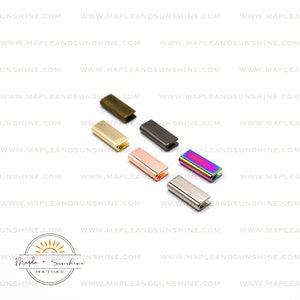 May include: Assortment of six metal slider buckles in various colors, including gold, silver, rose gold, black, rainbow, and bronze. The buckles are rectangular with rounded edges and a small opening on one side.