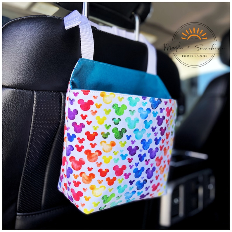 May include: A white car seat organiser with a rainbow Mickey Mouse print. The organiser has a teal blue top and a white strap with a hook for hanging on the back of a car seat. The text "Maple + Sunshine Boutique" is visible in the top right corner of the image.