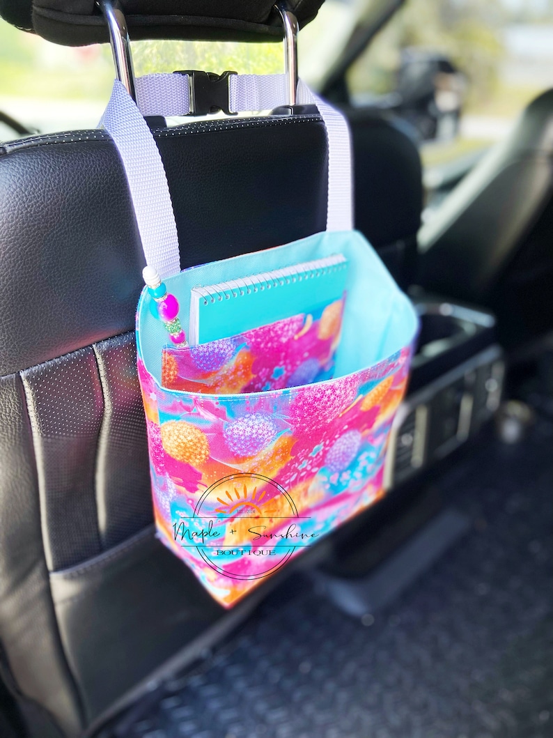 May include: A pink, orange and blue floral print car seat organiser with a white strap hanging from the headrest of a car seat. The organiser has a pocket for a notebook and a pen. The text "Maple + Sunshine Boutique" is printed on the organiser.