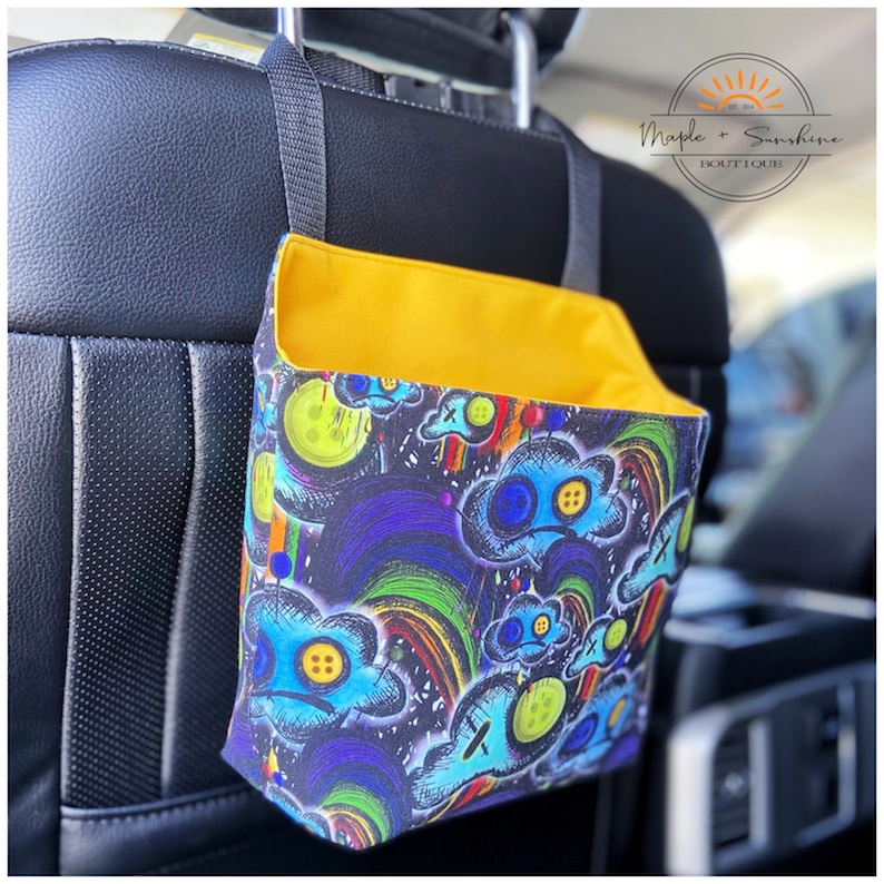 May include: A black car seat back with a yellow and black fabric hanging organiser. The organiser has a colourful design with clouds, rainbows, and buttons. The text "Maple + Sunshine Boutique" is visible in the top right corner of the image.