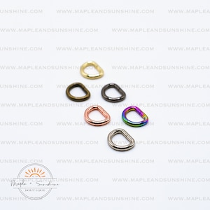 May include: Assortment of six metal D-rings in various colours, including gold, silver, black, rose gold, and rainbow. The D-rings are arranged on a white surface.