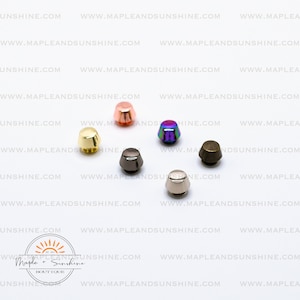 May include: Assortment of six metal cone-shaped rivets in various colors, including gold, silver, black, rose gold, iridescent, and bronze. The rivets are arranged on a white surface.