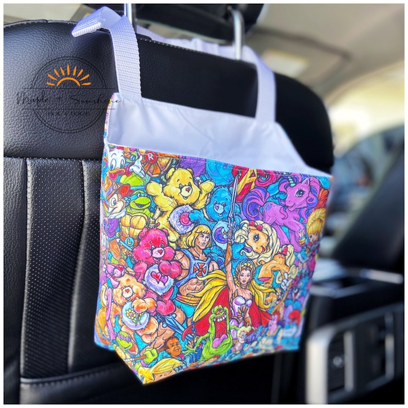 May include: A colourful car seat organiser with a white strap and hook. The organiser features a variety of cartoon characters including Care Bears, My Little Pony, He-Man, and Teenage Mutant Ninja Turtles.