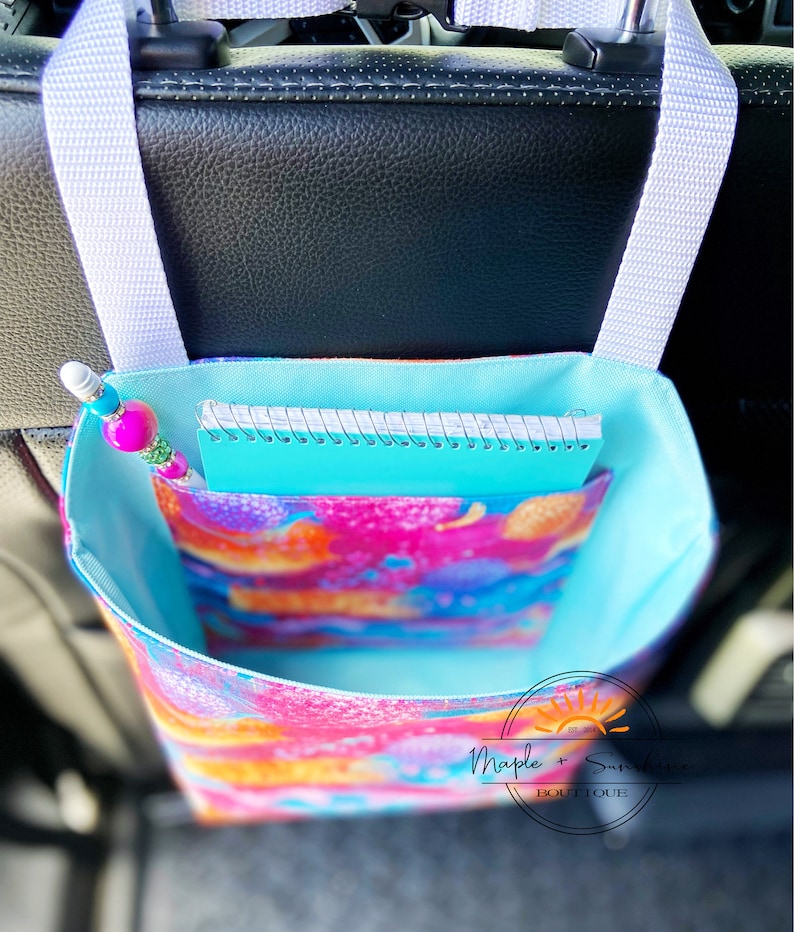May include: A turquoise and white fabric organiser with a colourful floral print hangs from the headrest of a car seat. The organiser has a pocket for a notebook and a pencil holder. The text "Maple + Sunshine Boutique" is visible on the organiser.