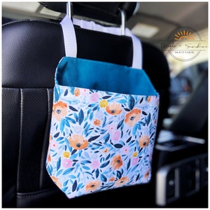 May include: A teal and white fabric car organiser with a floral print featuring orange, pink, and yellow flowers. The organiser hangs from the headrest of a car seat. The text "Maple + Sunshine Boutique" is visible on the organiser.