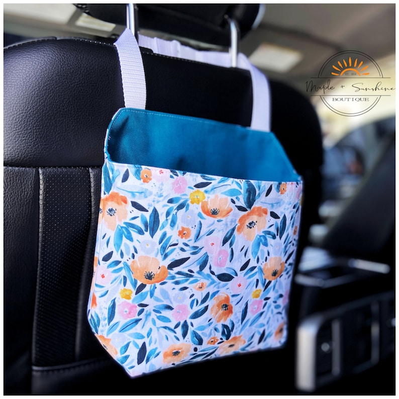 May include: A teal and white fabric car organiser with a floral print featuring orange, pink, and yellow flowers. The organiser hangs from the headrest of a car seat. The text "Maple + Sunshine Boutique" is visible on the organiser.