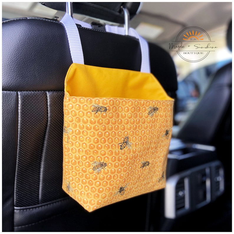 May include: Yellow car seat organiser with a honeycomb pattern and bees. The organiser has a white strap and hangs from the headrest. The text "Maple + Sunshine Boutique" is visible in the top right corner.