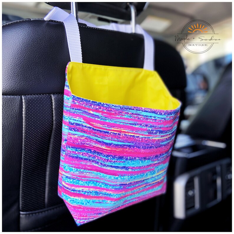 May include: A pink, blue and purple striped fabric car seat organiser with a yellow lining. The organiser is hanging from the headrest of a car seat. The text "Maple & Sunshine Boutique" is visible in the top right corner of the image.
