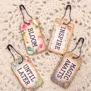 May include: Four decorative tags with black safety pins. Each tag has a floral or musical note background and a different word: BLOOM, INSPIRE, UNTIL LATER, and MAGIC AWAITS. The tags are rectangular with rounded corners.