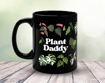 Può includere: Tazza in ceramica nera con una grafica bianca che dice "Plant Daddy" circondata da illustrazioni di piante verdi e rosa.