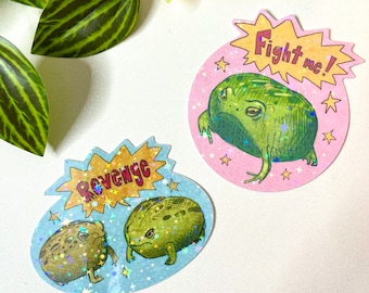 May include: Two stickers featuring cartoon frogs. One sticker has a green frog with the text "Fight me!" in a yellow burst. The other sticker has two brown frogs with the text "Revenge" in a yellow burst. Both stickers have a holographic glitter effect.
