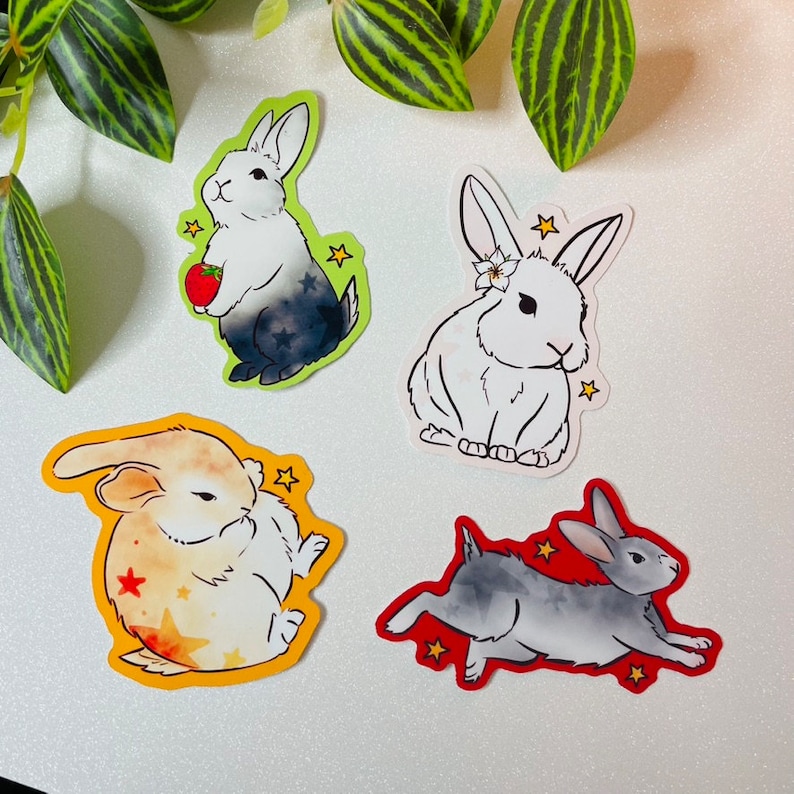 May include: Four sticker designs featuring different colored rabbits. The first rabbit is black and white with a strawberry in its paw. The second rabbit is white with a flower on its head. The third rabbit is yellow and white with a star on its chest. The fourth rabbit is gray with a star on its back.