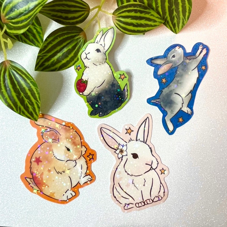 May include: Four colorful stickers featuring different cartoon bunnies. Each sticker has a sparkly, iridescent finish. The bunnies are in various poses, including holding a strawberry, sitting, and jumping.