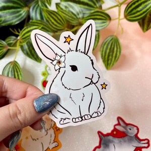 May include: A white rabbit sticker with a flower on its head and stars around it. The sticker is on a white background.