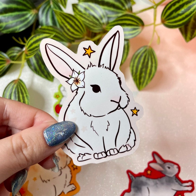 May include: A white rabbit sticker with a flower on its head and stars around it. The sticker is on a white background.