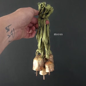 May include: A cluster of small, aged brass bells tied together with olive green fabric strips. The bells have a rustic, weathered appearance. The word "moss" is printed in white.