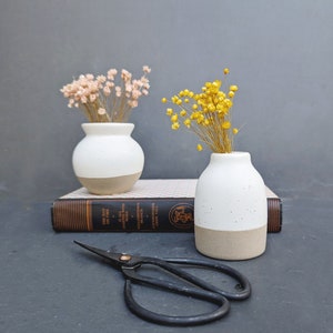 tiny bud vase with creamy white matte glazed 2/3 top and interior and a natural rough clay base for the bottom 1/3. light tan speckling throughout.