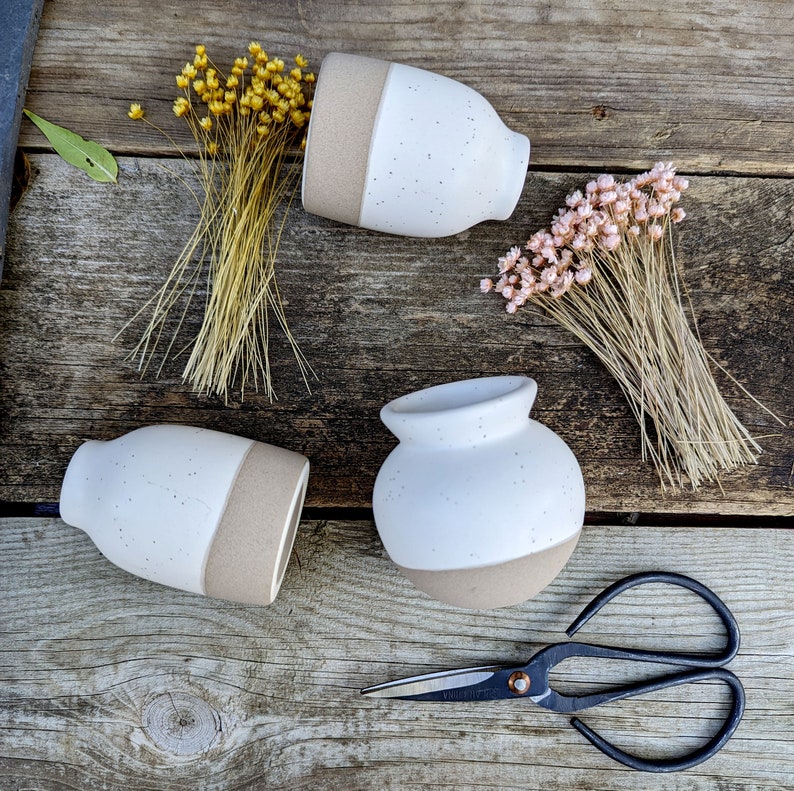 tiny bud vase with creamy white matte glazed 2/3 top and interior and a natural rough clay base for the bottom 1/3. light tan speckling throughout.