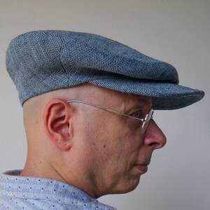 Puede incluir: Un hombre con una gorra plana de color azul con estampado y gafas. Lleva una camisa de botones azul claro con estampado blanco y azul.