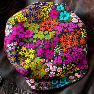 May include: A corduroy flat cap featuring a vibrant, multi-colored floral pattern on a black background. The fabric displays a dense arrangement of small flowers in shades of bright pink, yellow, orange, and turquoise, with a short, rounded brim.