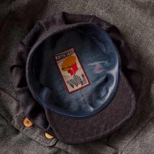 May include: A brown and blue patterned newsboy cap with a blue leather band. The cap has a label inside that reads "Built by Keith Loue, The Well Dressed Head".