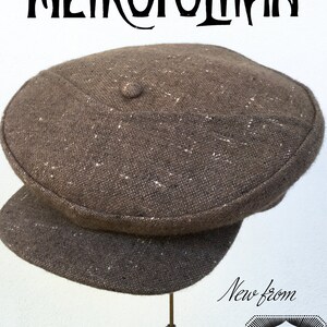 May include: A brown tweed newsboy cap with a button on top. The cap is on a stand and the text "The Metropolitan" is above the cap. The text "New from The Well Dressed Head" is below the cap.