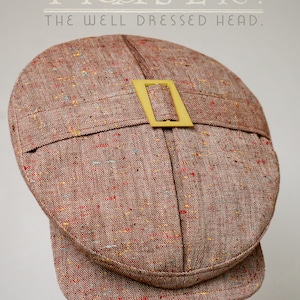 Puede incluir: Una gorra plana de tweed marrón con una hebilla dorada. La gorra tiene un patrón moteado y está hecha de una tela texturizada. El texto "The Fraser. The Well Dressed Head." es visible sobre la gorra.