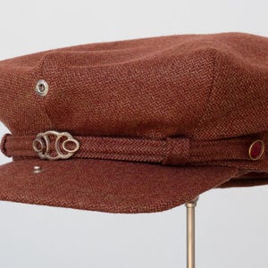 Puede incluir: Una gorra de newsboy de tweed marr&oacute;n con una hebilla de metal plateado y un bot&oacute;n rojo en el lateral.