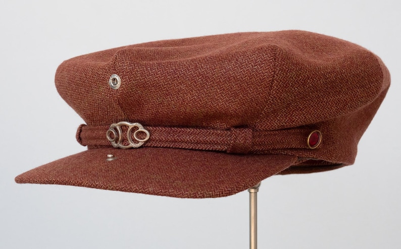 Puede incluir: Una gorra de newsboy de tweed marr&oacute;n con una hebilla de metal plateado y un bot&oacute;n rojo en el lateral.