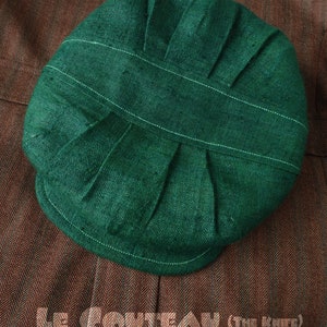 Puede incluir: Una gorra de lino verde oscuro con pliegues y un detalle de costura blanca. La gorra está sobre un tejido marrón con un patrón de espiga.