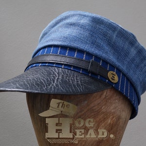 May include: A blue denim and black leather cap with a white and blue striped band. The cap has a brass button with the text "The Hog Head."
