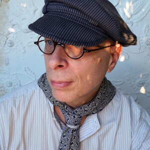 May include: A man wearing a striped, dark gray,  sailor-style cap with a black and white patterned scarf. He is wearing a white button-down shirt with thin, vertical stripes.