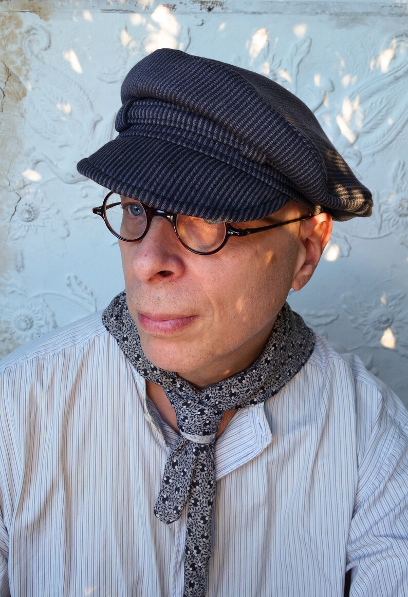 May include: A man wearing a striped, dark gray,  sailor-style cap with a black and white patterned scarf. He is wearing a white button-down shirt with thin, vertical stripes.