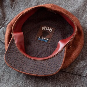 Puede incluir: Una gorra de newsboy de tweed marr&oacute;n con forro de cuero rojo. El interior de la gorra tiene una etiqueta que dice "WDH Caps. The True Made in Maine Cap.  For All Who Wear Caps."