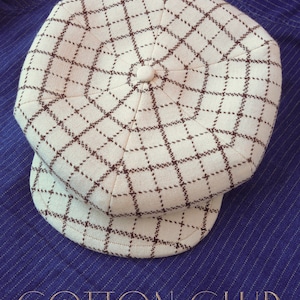 May include: A white and brown patterned newsboy cap with a brown brim. The cap has a grid pattern with brown lines on a white background. The cap is on a blue striped fabric.