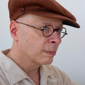 Puede incluir: Un hombre con una gorra de golf de tweed marr&oacute;n y gafas redondas de carey. Lleva una camisa de color beige claro con cuello.