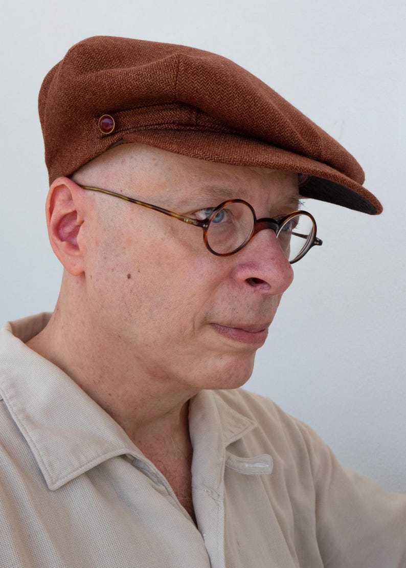 Puede incluir: Un hombre con una gorra de golf de tweed marr&oacute;n y gafas redondas de carey. Lleva una camisa de color beige claro con cuello.