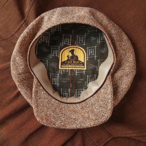 Puede incluir: Una gorra de newsboy de tweed marr&oacute;n y beige con una banda de cuero y una etiqueta que dice "K. Lo Bue for The Well Dressed Head Casquettes & Fine Sportswear Established 1914"