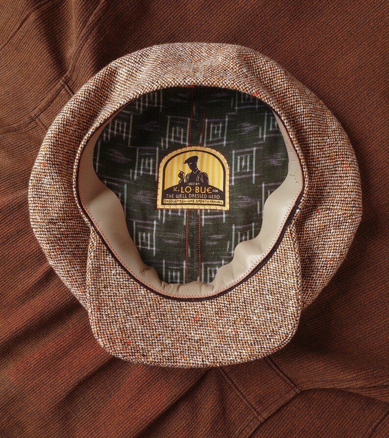 Puede incluir: Una gorra de newsboy de tweed marr&oacute;n y beige con una banda de cuero y una etiqueta que dice "K. Lo Bue for The Well Dressed Head Casquettes & Fine Sportswear Established 1914"