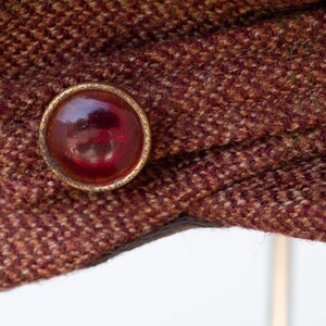 Puede incluir: Primer plano de una tela de tweed marr&oacute;n con un bot&oacute;n dorado con un centro de vidrio rojo.