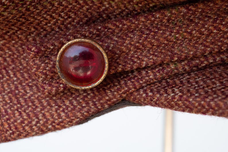 Puede incluir: Primer plano de una tela de tweed marr&oacute;n con un bot&oacute;n dorado con un centro de vidrio rojo.