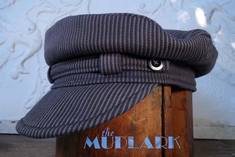 May include: A striped gray and black cotton cap with a button on the top. The cap has a wide brim and is folded down in the front. The cap is sitting on a wooden surface with the text "the MUDLARK" printed on it.