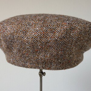 Puede incluir: Una gorra de newsboy de tweed marr&oacute;n, blanco y naranja con una corona redonda y una visera plana.