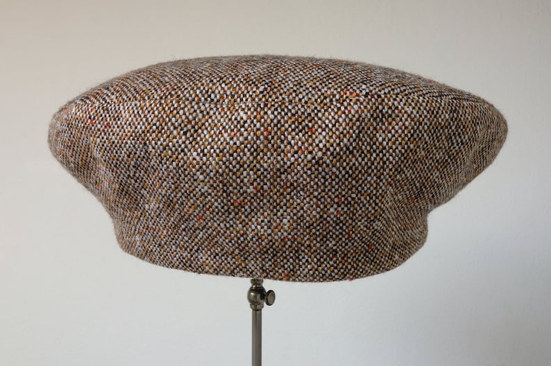 Puede incluir: Una gorra de newsboy de tweed marr&oacute;n, blanco y naranja con una corona redonda y una visera plana.