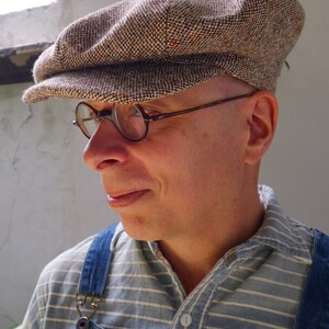 Puede incluir: Un hombre con una gorra de newsboy de tweed marr&oacute;n y beige con una visera marr&oacute;n. Tambi&eacute;n lleva gafas redondas y una camisa a rayas azules y blancas con un mono vaquero.