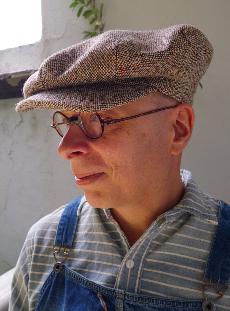 Puede incluir: Un hombre con una gorra de newsboy de tweed marr&oacute;n y beige con una visera marr&oacute;n. Tambi&eacute;n lleva gafas redondas y una camisa a rayas azules y blancas con un mono vaquero.