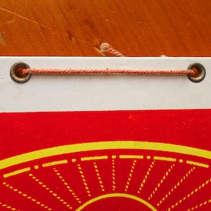 May include: A white and red calendar with a yellow sun design. The calendar is hanging from a string with two metal eyelets.