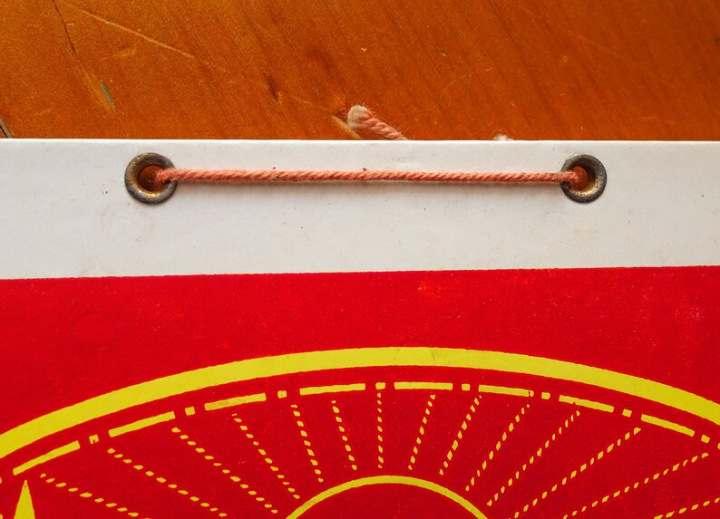 May include: A white and red calendar with a yellow sun design. The calendar is hanging from a string with two metal eyelets.